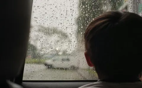 Dziecko patrzy przez zaparowane kroplami deszczu okno, szukając pomysłów na zabawy na deszczowe dni.