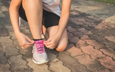 Kobieta w sportowym stroju klęczy i zawiązuje sznurówki w butach do **slow joggingu**.