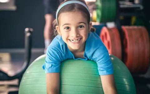 Uśmiechnięta dziewczynka na zielonej piłce gimnastycznej. W tle sztangi, idealne zajęcia sportowe dla 3 latka.