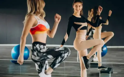 Dwie kobiety ćwiczą na stepach. Jedna w kolorowym topie i wzorzystych legginsach, druga w czarnym body i beżowych legginsach. TBC co to znaczy?