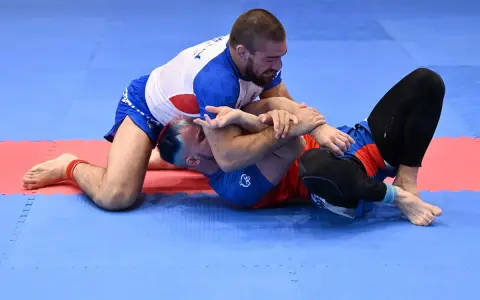 Dwóch mężczyzn walczy w brazylijskim jiu jitsu na niebiesko-czerwonej macie. Jeden z nich klęczy, drugi leży.
