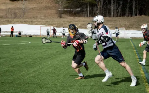 Dwóch zawodników w akcji podczas meczu lacrosse. Jeden biegnie z pałką, drugi go ściga.
