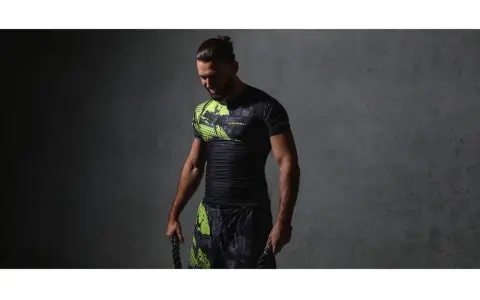 Mężczyzna w czarnym rashguardzie z neonowymi wstawkami i spodenkami, trzymający liny treningowe. Rashguard co to? Odzież sportowa.