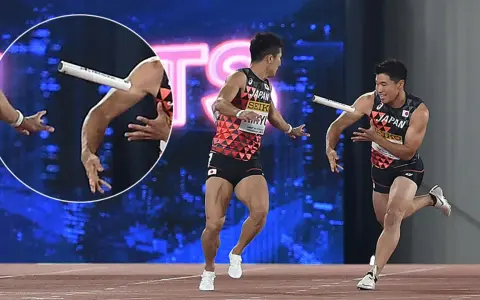 Dwóch lekkoatletów z Japonii w trakcie biegu z pałeczką. Jeden z nich biegnie, drugi wyciąga rękę po pałeczkę.