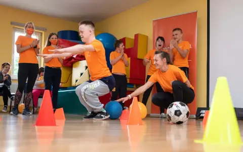 Wesołe wf dla dzieci z piłką i pachołkami. Uśmiechnięci uczestnicy i trenerzy w pomarańczowych koszulkach.