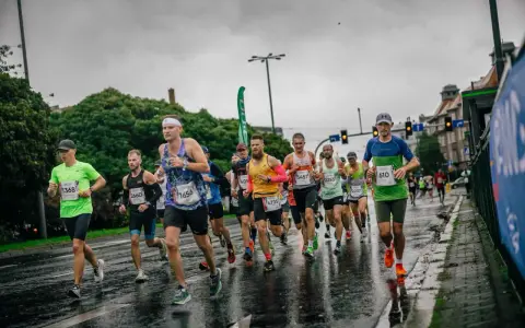 Zawodnicy biegną w deszczu podczas maratonu. Mokra nawierzchnia odbija światła, a biegacze w kolorowych strojach walczą o jak najlepszy czas.