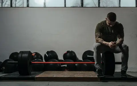 Mężczyzna z tatuażami odpoczywa po treningu crossfit w domu, otoczony piłkami lekarskimi i sztangą.