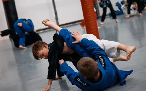 Dzieci ćwiczą jujitsu, walcząc na macie w niebieskich i czarnych kimonach.