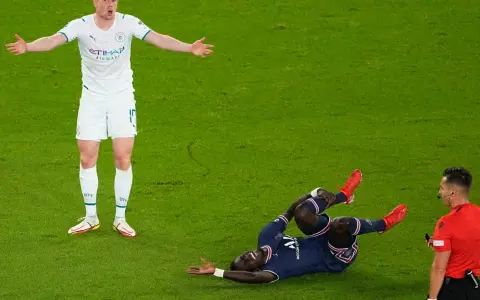 Piłkarz leży na trawie po brutalnym faulu, drugi zawodnik z niedowierzaniem rozkłada ręce, a sędzia obserwuje zdarzenie.