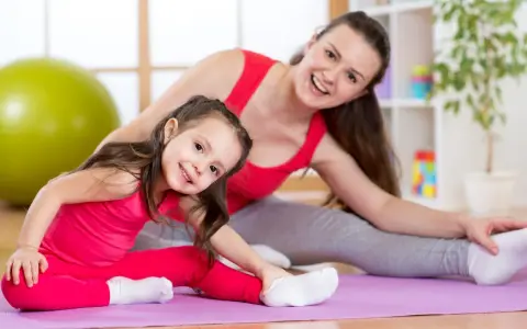 Mama i córka wykonują ćwiczenia rozciągające dla dzieci na macie. Uśmiechnięte, w różowych i szarych strojach.