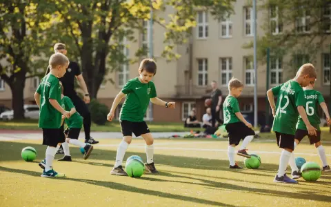 Dzieci ćwiczą na boisku w ramach szkółki piłkarskiej. W zielonych strojach biegają z piłkami, ucząc się gry.