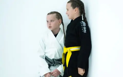 Dwie dziewczynki w kimonach do BJJ, jedna w białym z szarym pasem, druga w czarnym z żółtym pasem. Ich pasy bjj dzieci to symbol postępów w treningach.