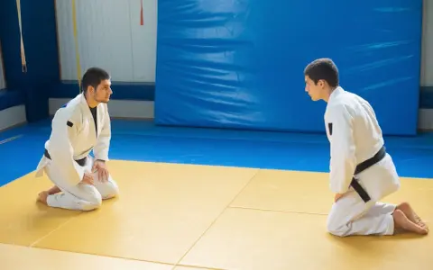 Dwóch judoków w białych kimonach, jeden z czarnym pasem judo, kłania się sobie na macie.