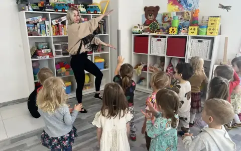 Wesołe zajęcia rytmiczne w przedszkolu. Nauczycielka tańczy z dziećmi wśród zabawek na półkach.