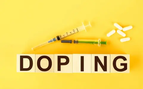 Dwa strzykawki i białe kapsułki na żółtym tle. Słowo "DOPING" napisane na drewnianych klockach.