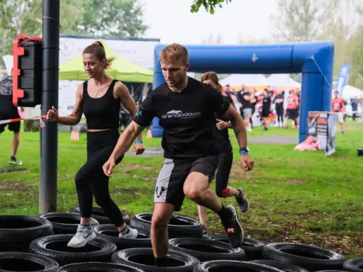 Zawodnicy pokonują tor przeszkód z opon. Kobieta i mężczyzna biegną, a za nimi inni uczestnicy. To prawdziwa survival race!