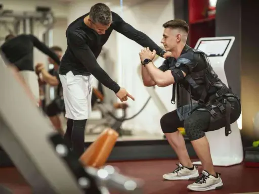 Trening EMS: Instruktor koryguje pozycję mężczyzny w kamizelce stymulującej mięśnie podczas przysiadu.