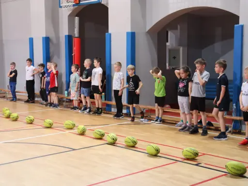 Dzieci w hali sportowej czekają na rozpoczęcie gry i zabawy. Na parkiecie leżą żółte piłki.