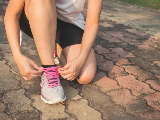 Kobieta w sportowym stroju klęczy i zawiązuje sznurówki w butach do **slow joggingu**.