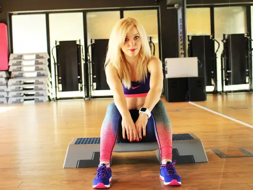 Blondynka w sportowym stroju siedzi na stopniu, gotowa do ćwiczeń na brzuch.