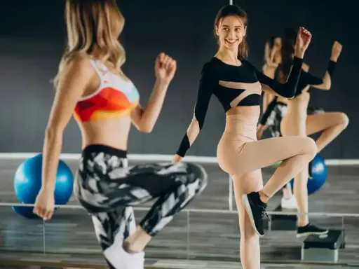 Dwie kobiety ćwiczą na stepach. Jedna w kolorowym topie i wzorzystych legginsach, druga w czarnym body i beżowych legginsach. TBC co to znaczy?
