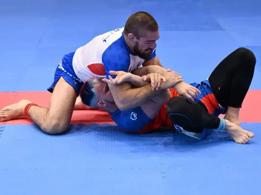 Dwóch mężczyzn walczy w brazylijskim jiu jitsu na niebiesko-czerwonej macie. Jeden z nich klęczy, drugi leży.