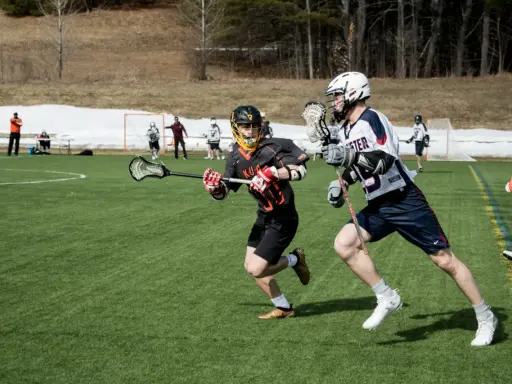 Dwóch zawodników w akcji podczas meczu lacrosse. Jeden biegnie z pałką, drugi go ściga.