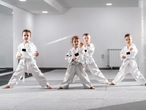Cztery dzieci w białych kimonach ćwiczą judo dla dzieci. Stoją w pozycji bojowej na macie w jasnej sali treningowej.