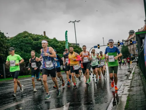 Zawodnicy biegną w deszczu podczas maratonu. Mokra nawierzchnia odbija światła, a biegacze w kolorowych strojach walczą o jak najlepszy czas.