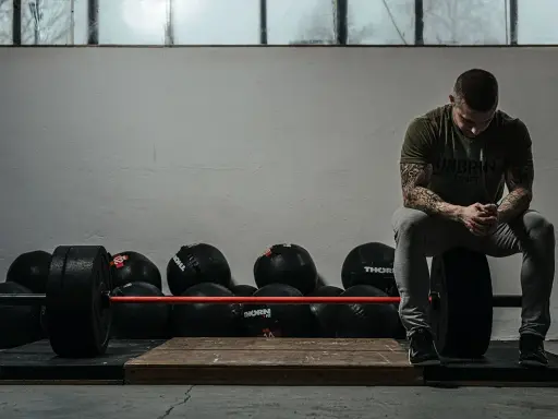 Mężczyzna z tatuażami odpoczywa po treningu crossfit w domu, otoczony piłkami lekarskimi i sztangą.