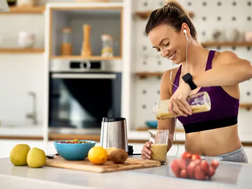 Uśmiechnięta kobieta przygotowuje smoothie, idealne jako co zjesc przed biegiem. Na blacie owoce i blender.
