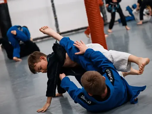 Dzieci ćwiczą jujitsu, walcząc na macie w niebieskich i czarnych kimonach.