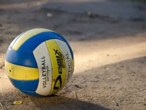 Piłka do siatkówki plażowej, niebiesko-żółta, leży na piasku. Różni się od halowej miękkością i wagą.