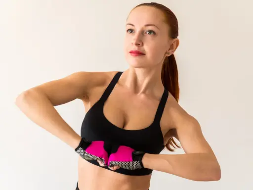 Kobieta w sportowym topie i rękawiczkach wykonuje ćwiczenia na biust, napinając mięśnie klatki piersiowej.