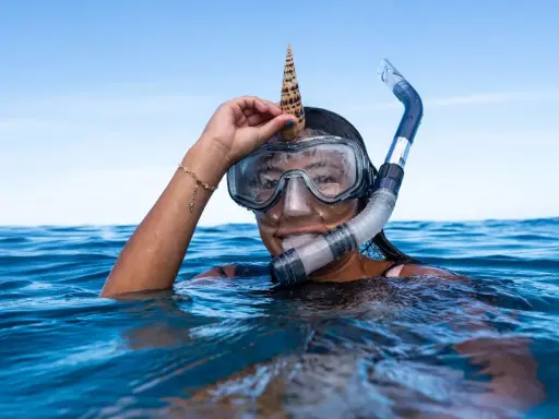 Dziewczyna w masce i fajce do snorkelingu trzyma muszlę na głowie. To właśnie snorkeling!