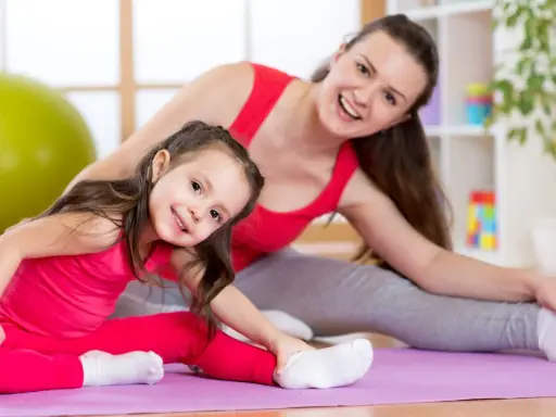Mama i córka wykonują ćwiczenia rozciągające dla dzieci na macie. Uśmiechnięte, w różowych i szarych strojach.