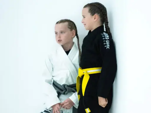 Dwie dziewczynki w kimonach do BJJ, jedna w białym z szarym pasem, druga w czarnym z żółtym pasem. Ich pasy bjj dzieci to symbol postępów w treningach.