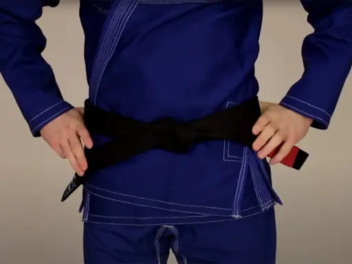 Osoba w niebieskim gi demonstruje wiązanie pasa bjj. Czarny pas jest ciasno zawiązany na wysokości talii.
