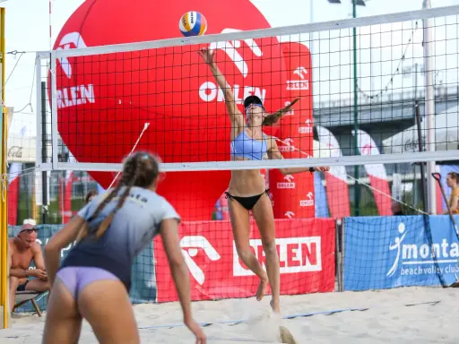 Dwie zawodniczki grają w siatkówkę plażową. Jedna z nich właśnie serwuje piłkę, druga jest w gotowości do obrony.