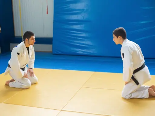 Dwóch judoków w białych kimonach, jeden z czarnym pasem judo, kłania się sobie na macie.