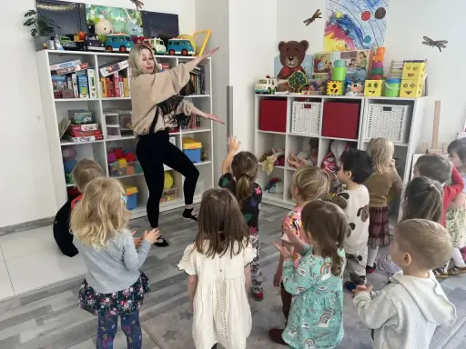 Wesołe zajęcia rytmiczne w przedszkolu. Nauczycielka tańczy z dziećmi wśród zabawek na półkach.