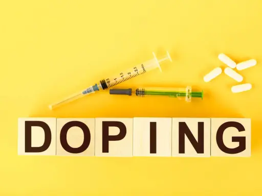 Dwa strzykawki i białe kapsułki na żółtym tle. Słowo "DOPING" napisane na drewnianych klockach.