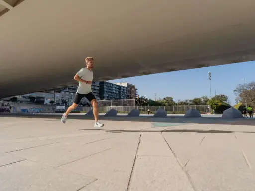 Mężczyzna w lekkim stroju sportowym biegnie po betonowym placu, przygotowując się do biegu na 10 km. W tle budynki i drzewa.