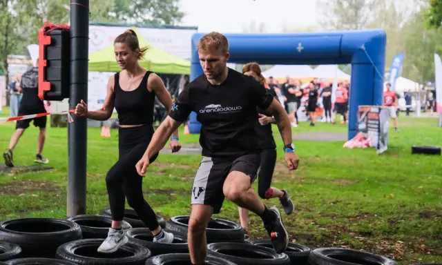 Zawodnicy pokonują tor przeszkód z opon. Kobieta i mężczyzna biegną, a za nimi inni uczestnicy. To prawdziwa survival race!