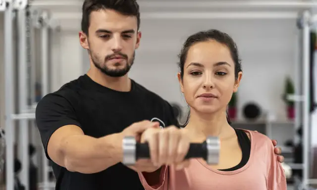 Trening anaerobowy z trenerem. Kobieta ćwiczy z hantlem, mężczyzna koryguje jej ruchy.