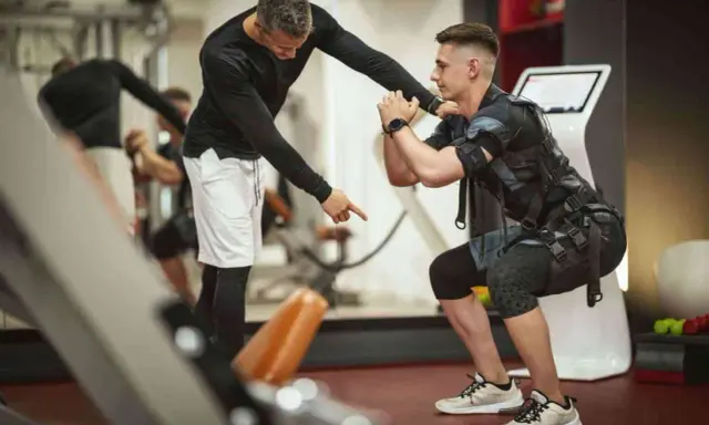 Trening EMS: Instruktor koryguje pozycję mężczyzny w kamizelce stymulującej mięśnie podczas przysiadu.