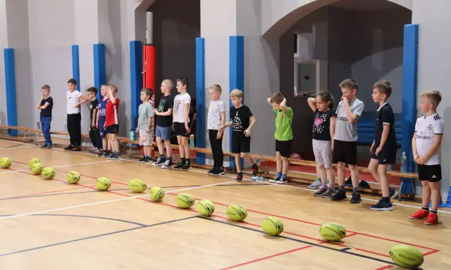 Dzieci w hali sportowej czekają na rozpoczęcie gry i zabawy. Na parkiecie leżą żółte piłki.