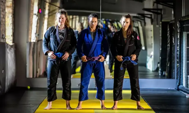 Trzy kobiety w kimonach do BJJ. Środkowa ma purpurowy pas bjj, a pozostałe niebieskie.