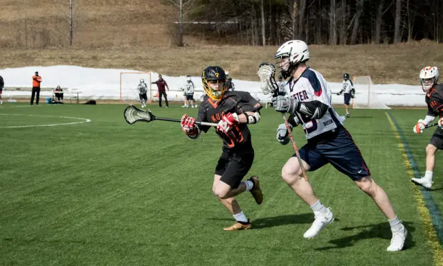 Dwóch zawodników w akcji podczas meczu lacrosse. Jeden biegnie z pałką, drugi go ściga.