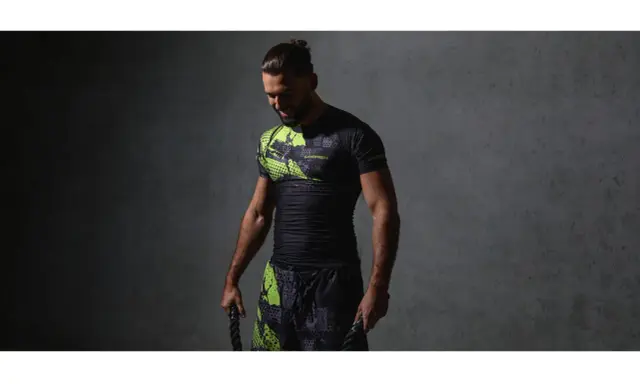 Mężczyzna w czarnym rashguardzie z neonowymi wstawkami i spodenkami, trzymający liny treningowe. Rashguard co to? Odzież sportowa.